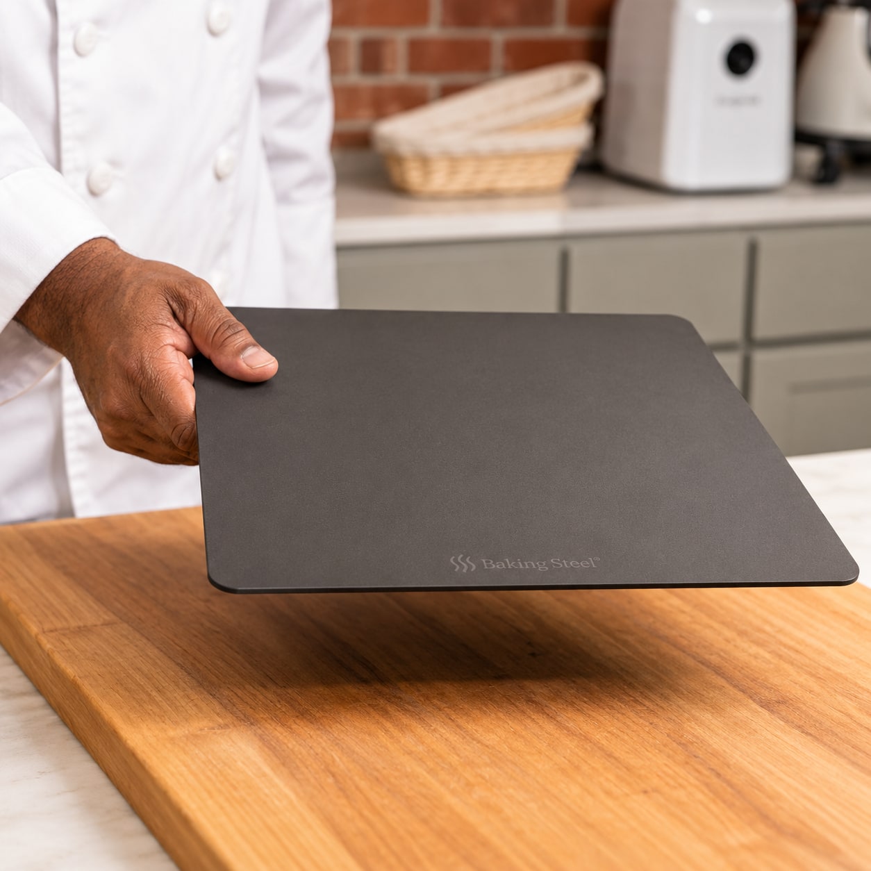 A baker holding a rectangular Baking Steel plate above a wooden work surface